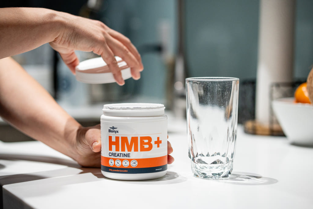 Person holding a container of Blonyx HMB+ Creatine with a glass on a kitchen counter.