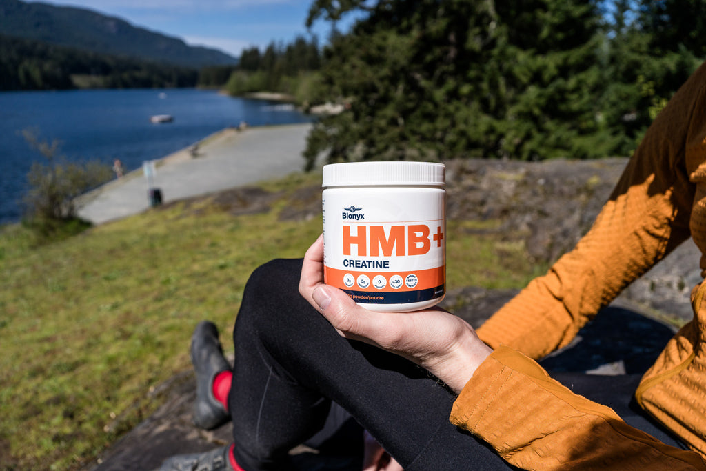 Person holding a HMB+ Creatine supplement container outdoors by a lake