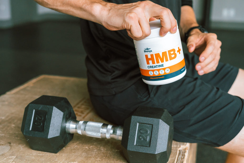 Person holding a supplement container labeled HMB+ Creatine with a dumbbell beside them