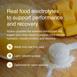 Hydra+ real food electrolytes powder shown in a white scoop on a wooden surface, with a glass of yellow drink and cubed mango in the background. Text overlays describe the product benefits.