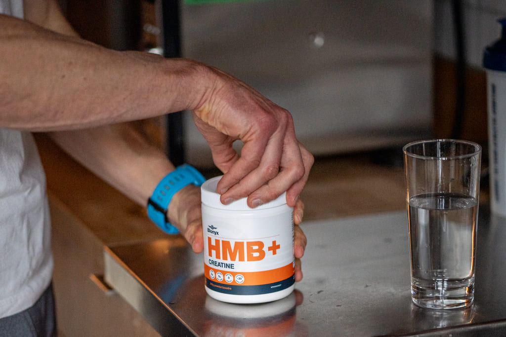 Person holding a container of HMB+ Creatine supplement with a glass of water on a counter.